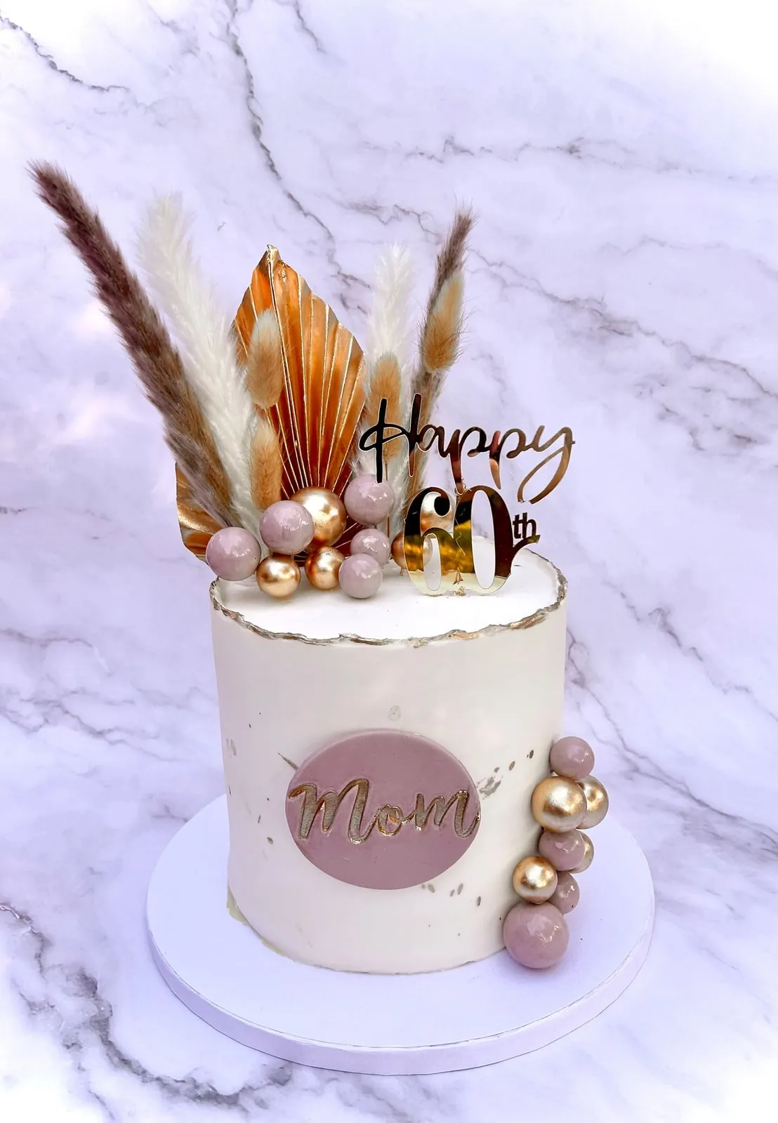 Ivory sixtieth birthday cake with pampas grass, gold spheres and a plaque